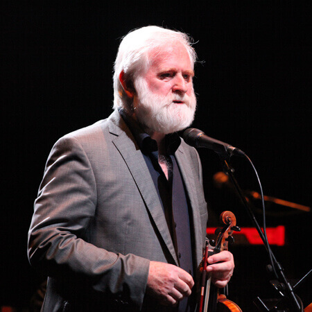 the white-haired, white-bearded john sheahan is lit up on a dark stage