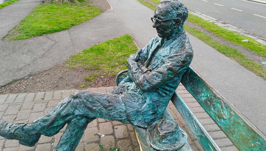 a sculpture of patrick kavanagh sitting on a full-sized bench with his arms folded