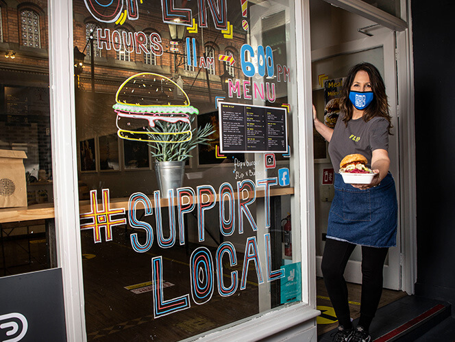 Food server in mask stands outside by a window display that says support local