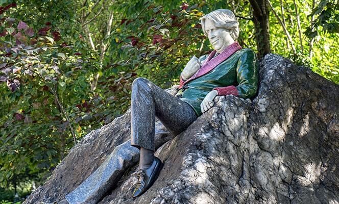 sculpture of oscar wilde leaning back on a rock wearing a green and red jacket with black shoes